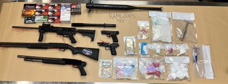Items including long-guns, pistols, cash, and suspected packaged drugs are displayed as evidence on a table. 