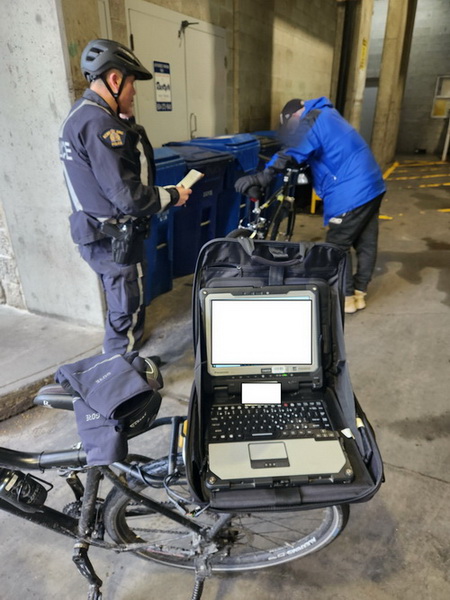 Police officer standing next to his bicycle with a police laptop open. He is talking into his radio and looking at a male dressed in a blue jacket and black pants who is leaning against a black bicycle