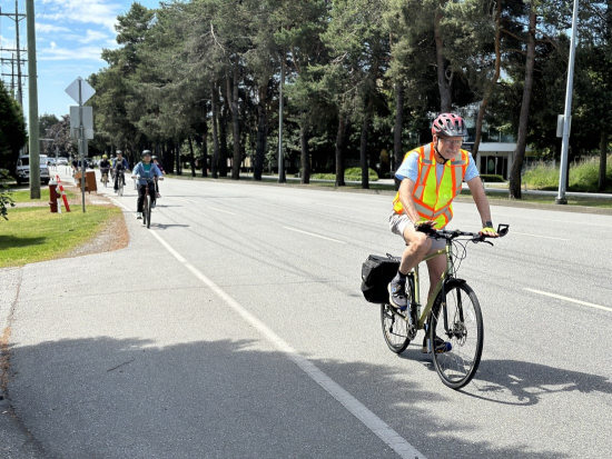 Bénévole en vélo avec un gilet de sécurité et un casque qui roule du côté droit de la route et qui est suivi d’autres cyclistes.