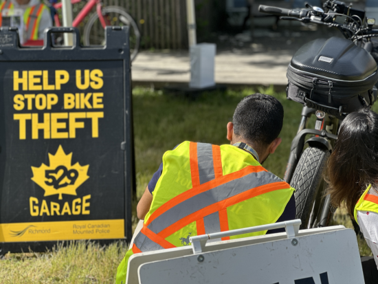 volunteer in a safety vest inspecting a bicycle. Sign on the ground reads: <q>Help us stop bike theft. 529 Garage.</q>