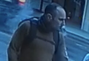 A balding man with a long sleeved brown shirt wearing a backpack