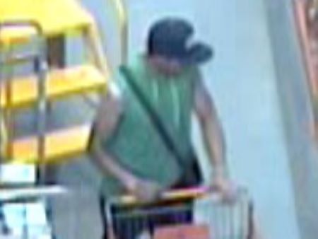 Un homme portant un haut vert sans manches, un pantalon noir et une casquette de baseball bleue se trouve debout dans l’allée d’un commerce et tient un panier de magasinage d’une main. 