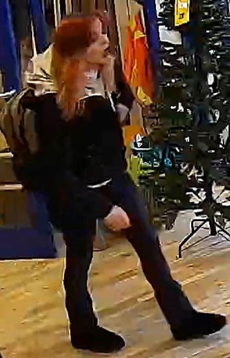 A woman with red hair, dark jacket and pants, and a white sweater, walks by a Christmas tree for sale in a retail store. 
