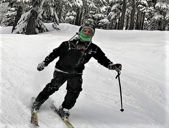 Le suspect qui porte un manteau et un pantalon de ski foncés, un casque de ski ainsi qu’un cache-cou vert vif. L’homme est sur une pente de ski alpin et tient des bâtons de ski.
