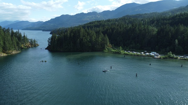 An aerial photograph of the BC RCMP Zodiac boat in Harrison Lake at Ten Mile Bay campsite. Campers inside vehicles and trailers are parked at the waterfront.