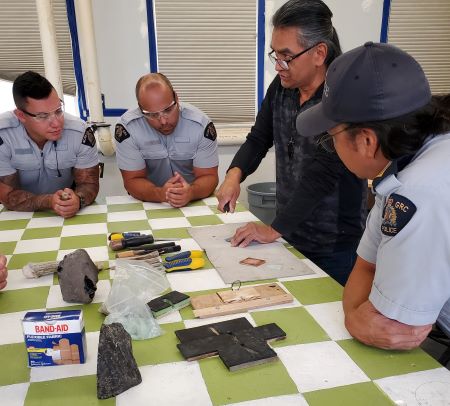 Démonstration : Des policiers en uniforme sont rassemblés autour d’Ed Jensen pendant qu’il fait une démonstration de technique de taille de silex sur une table à carreaux verts et blancs. Des bandages, des outils et d’autres fournitures sont posés sur la table. 