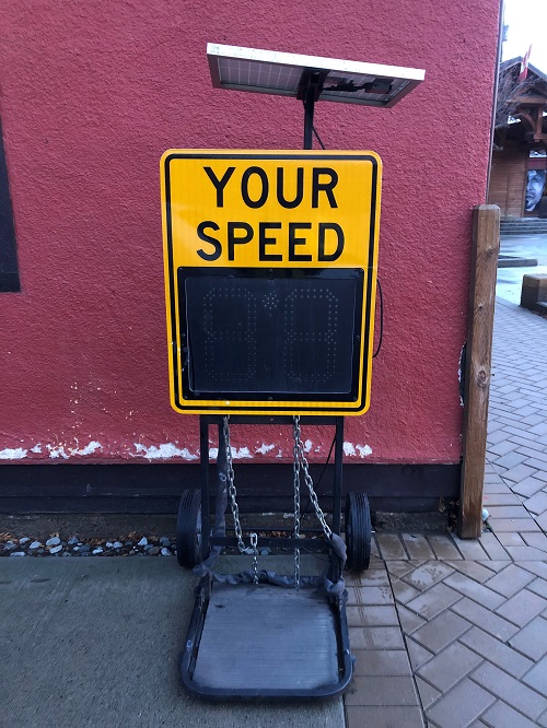 Yellow speed sign with a solar panel on top, sitting on the sidewalk in front of the Merritt Community Police Office.